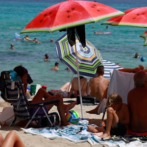 Auf dem Symbolfoto von August 2022 sieht man Strand, Meer und Menschen. Sie schwimmen, liegen in der Sonne oder unter Sonnenschirmen im Schatten.