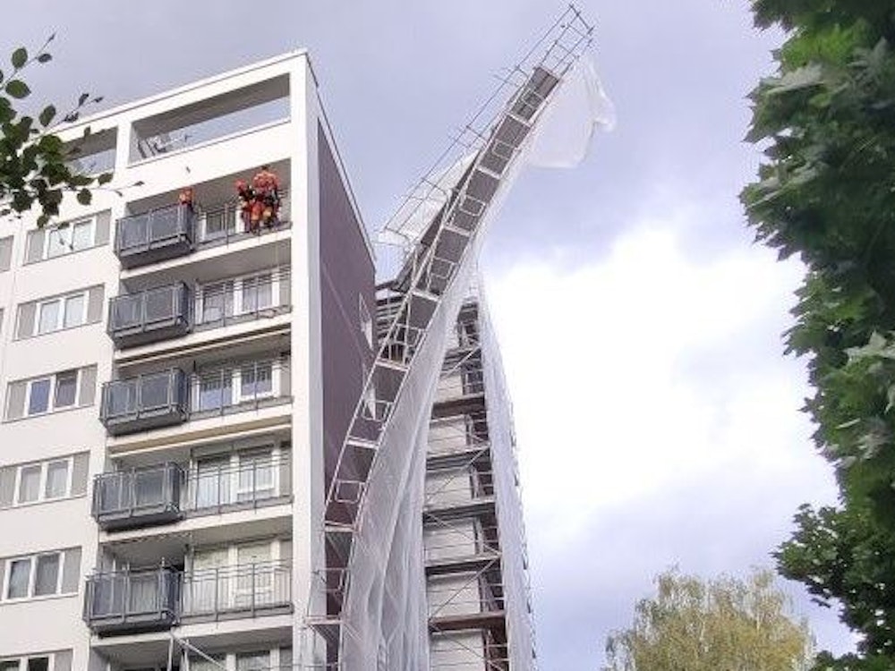 Ein Baugerüst hat sich von der Fassade eines Mehrfamilienhauses gelöst und droht umzukippen. Auf einem Balkon stehen mehrere Einsatzkräfte der Höhenrettung.