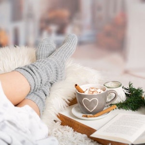 Frau liegt eingekuschelt mit dicken Socken, heißer Schokolade und einem Buch auf dem Sofa.