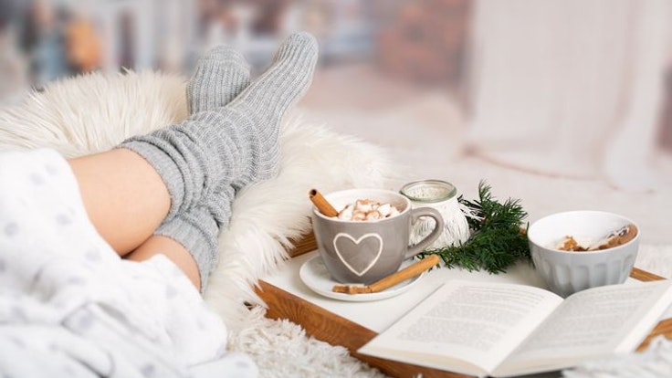 Frau liegt eingekuschelt mit dicken Socken, heißer Schokolade und einem Buch auf dem Sofa.