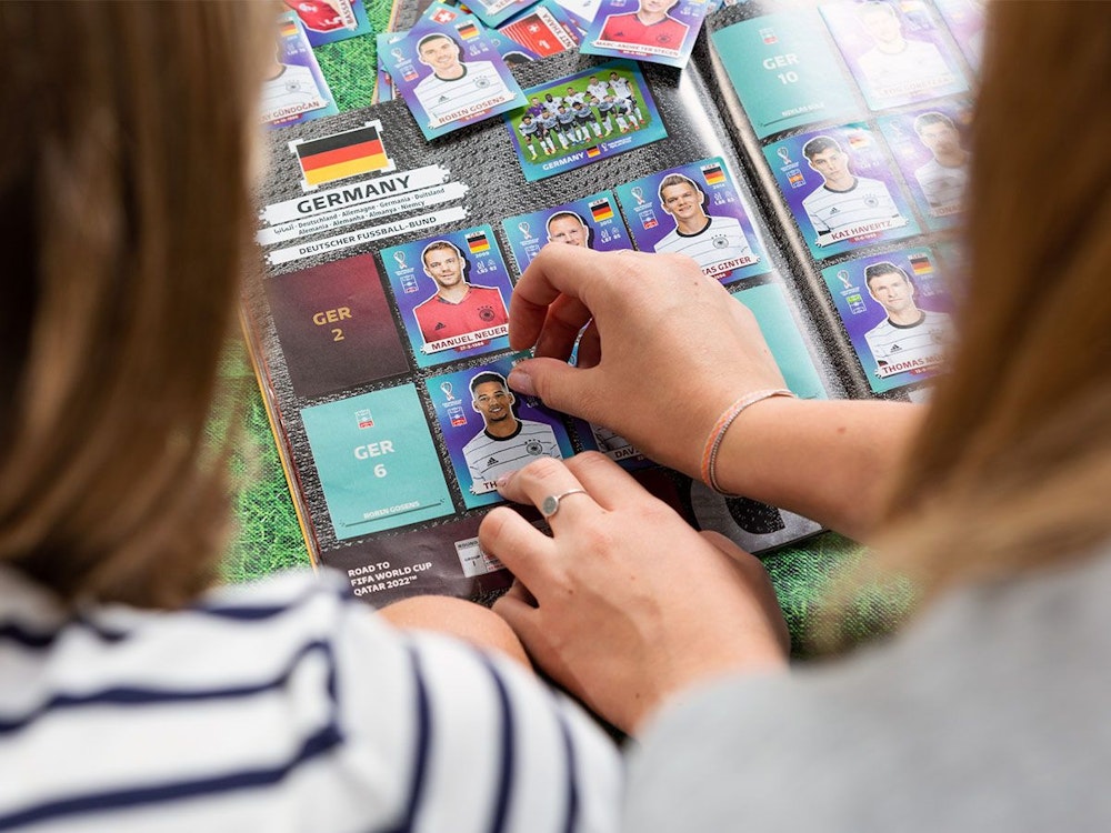 Zwei Mädchen sitzen am Tisch und kleben Panini-WM-Sticker der deutschen Fußball-Nationalmannschaft in das FIFA World Cup 2022 Qatar Stickeralbum ein.
