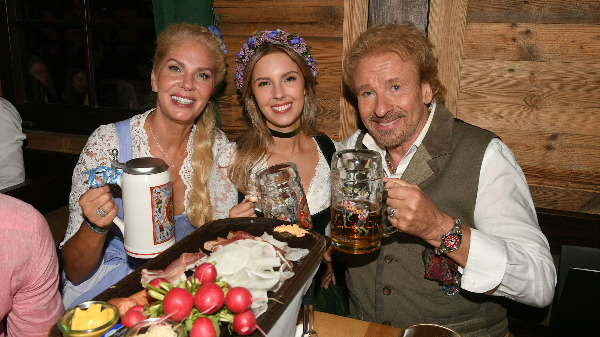 Auf dem Bild sitzen Karina Mroß, Melinda Mroß und Thomas Gottschalk vor einer Brotzeit-Platte. Alle drei posieren mit Bierkrug in der Hand und lächeln in die Kamera.