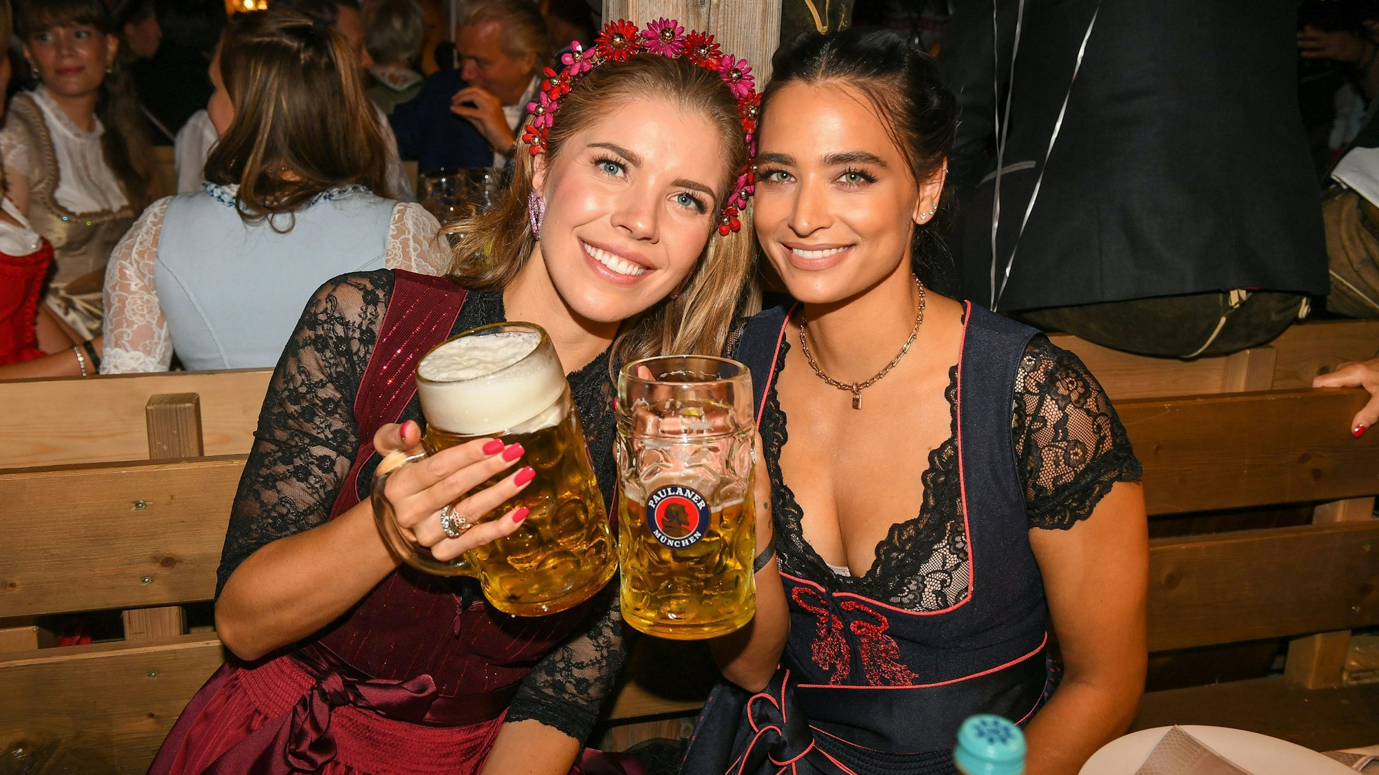 Victoria Swarovski und Amira Pocher halten beide Bierkrüge in der Hand und lächeln in die Kamera. Beide Frauen haben sich für eine schwarz-rote Dirndl-Kombination entschieden.
