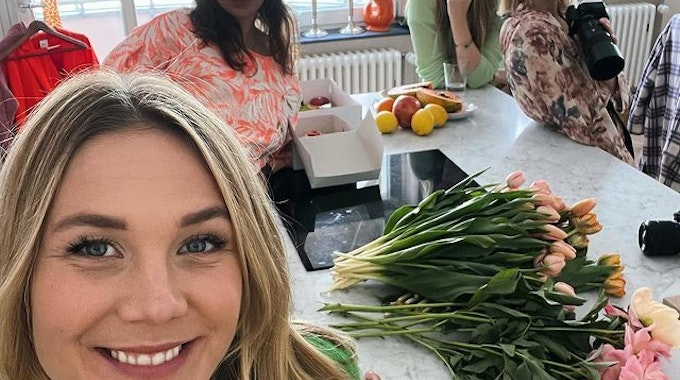 Alina Merkau macht ein Selfie. Im Hintergrund stehen drei weitere Frauen. Hinter ihr auf der Küchenarbeitsplatte liegen verschiedene Blumen.