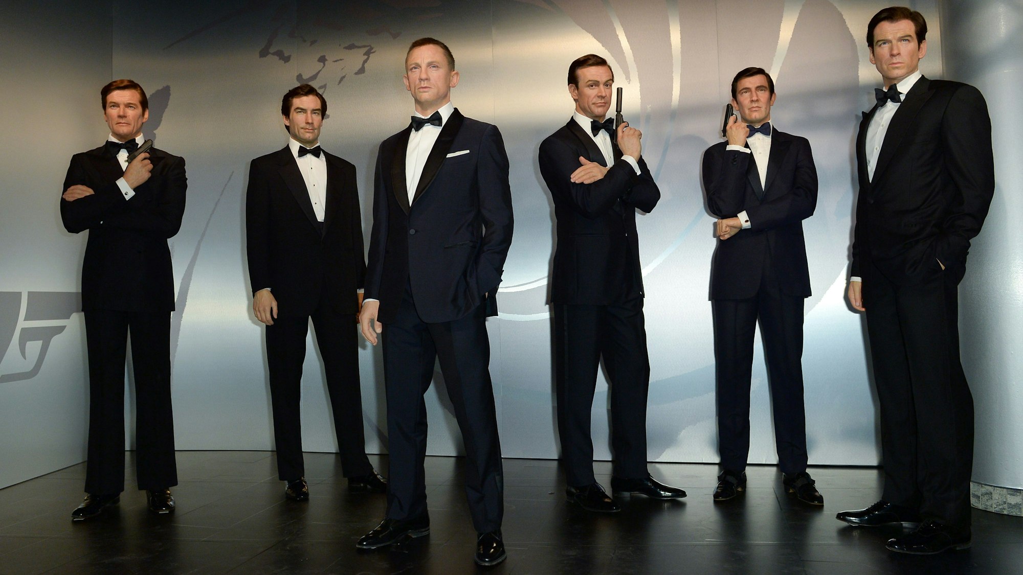 Die sechs Bond-Darsteller sind als Wachsfiguren ausgestellt. Jede Figur trägt einen schwarzen Anzug, ein weißes Hemd und eine schwarze Fliege. Drei Figuren halten zusätzlich noch eine Pistole in der Hand.