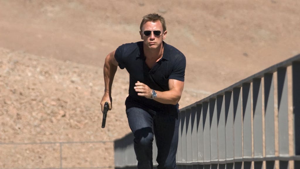 Daniel Craig rennt in der Szene des Films „Ein Quantum Trost“ einen Weg entlang. In der rechten Hand hält er eine Pistole. Er trägt Jeans, ein schwarzes T-Shirt und eine dunkle Sonnenbrille.