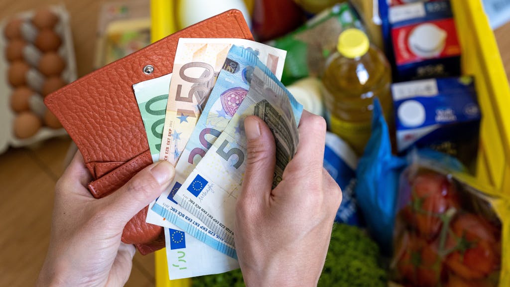 Eine Einkaufskiste mit Lebensmitteln steht auf einem Küchentisch, während eine Frau Euro-Banknoten in den Händen hält. Ab Oktober ändert sich einiges, auch finanziell.
