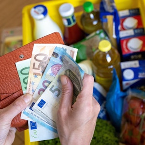 Eine Einkaufskiste mit Lebensmitteln steht auf einem Küchentisch, während eine Frau Euro-Banknoten in den Händen hält. Ab Oktober ändert sich einiges, auch finanziell.
