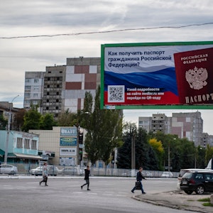 Ukraine, Luhansk: Menschen überqueren eine Straße mit einem Plakat mit der Aufschrift ‚Wie man den Pass eines russischen Bürgers bekommt‘.