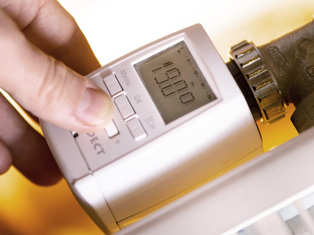 Eine Hand fasst an ein modernes Thermostat mit digitaler Anzeige an einer Heizung, wobei 19. Grad Celsius auf einem kleinen Display angezeigt werden.