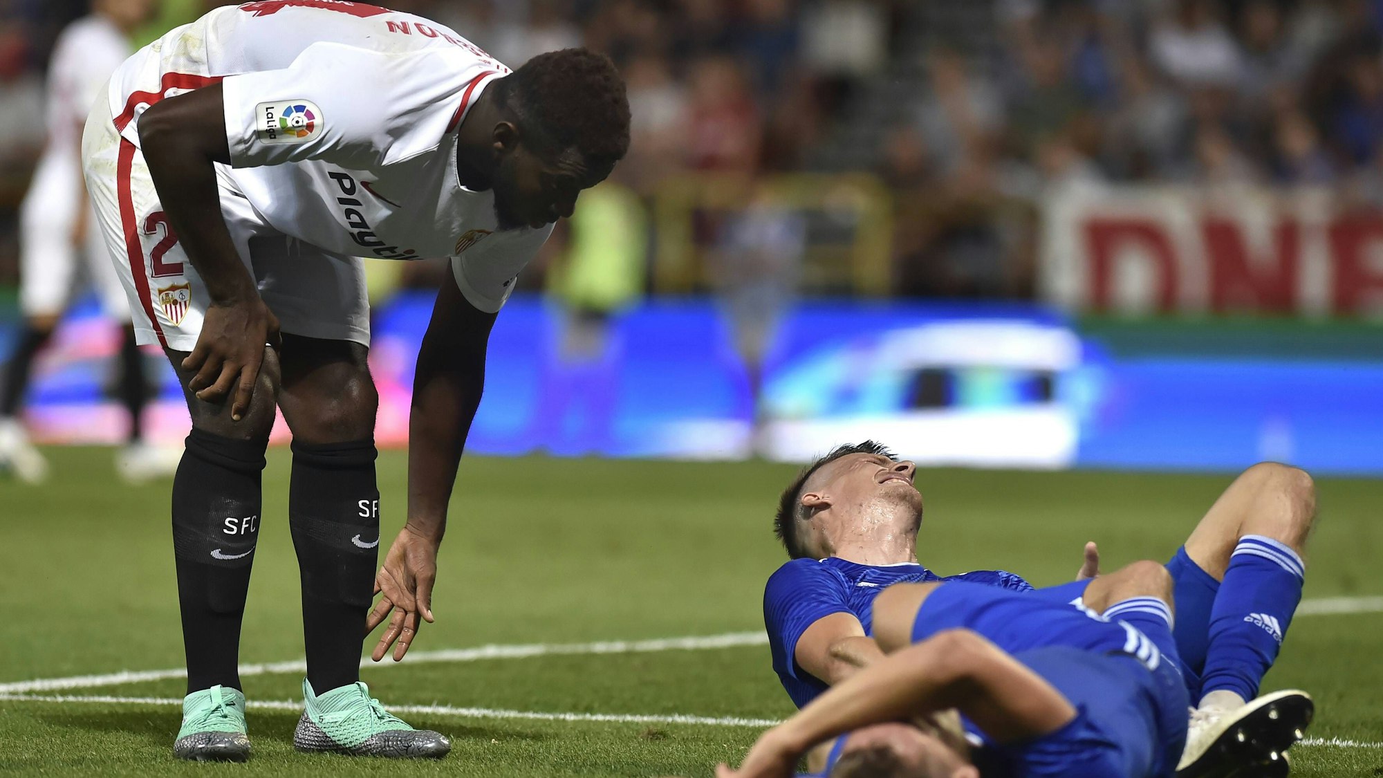 Joris Gnagnon, damals noch im Trikot des FC Sevilla, bückt sich, während zwei Gegenspieler verletzt auf dem Rasen liegen.