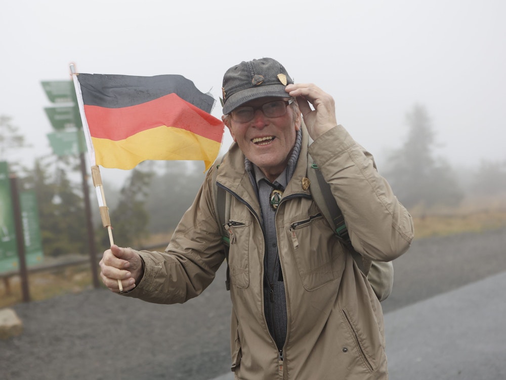 Extremwanderer Benno Schmidt, besser bekannt als „Brocken-Benno“, steht am Rande der Feierstunde zum Tag der Deutschen Einheit auf dem Brocken und hält eine Deutschland-Fahne in der Hand.