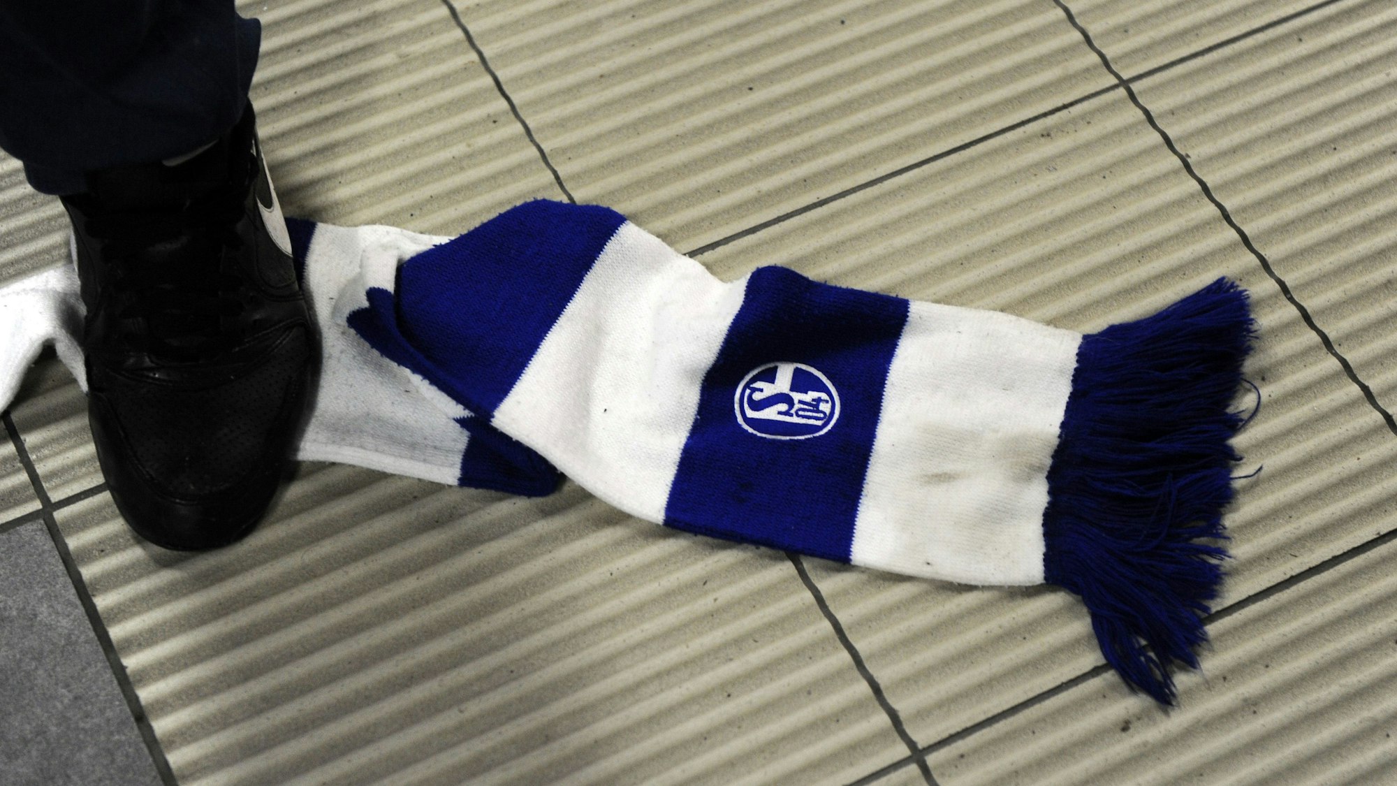 Ein Fanschal von Schalke 04 liegt auf dem Boden, eine Person tritt mit einem Fuß darauf.