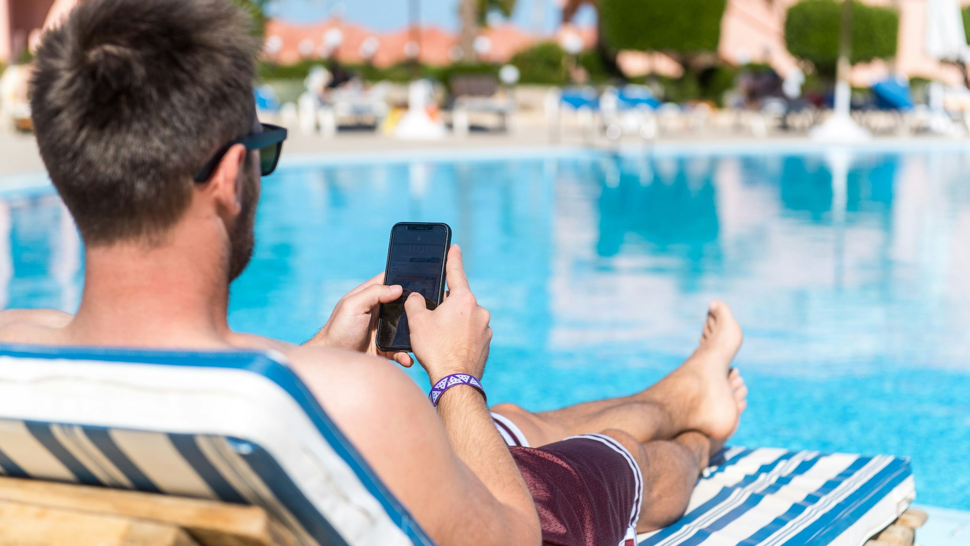 Ein Mann sitzt in der Sonne an einem Pool und hat sein Smartphone in der Hand