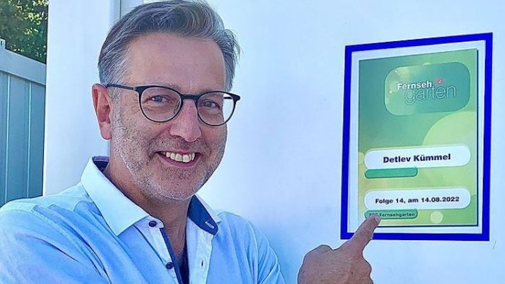 Detlev Kümmel lacht auf einem Foto fröhlich in die Kamera und zeigt auf eine Werbetafel des ZDF-„Fernsehgarten“.