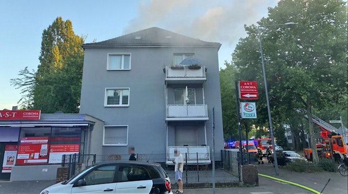 Qualm steigt aus einer Mehrfamilienhaus an einer Straße in Köln. Vor dem Haus steht die Feuerwehr.