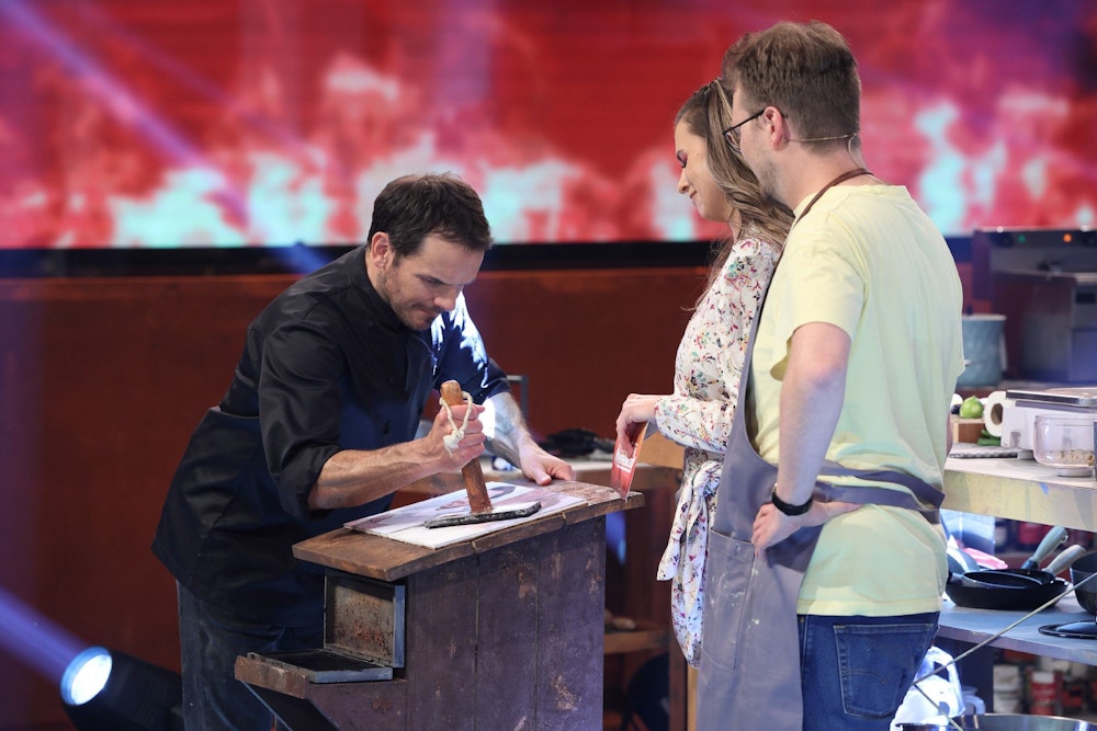 Steffen Henssler demonstriert Laura Wontorra und Maxi Gstettenbauer, wie er etwas an einem Tisch zerstampft.