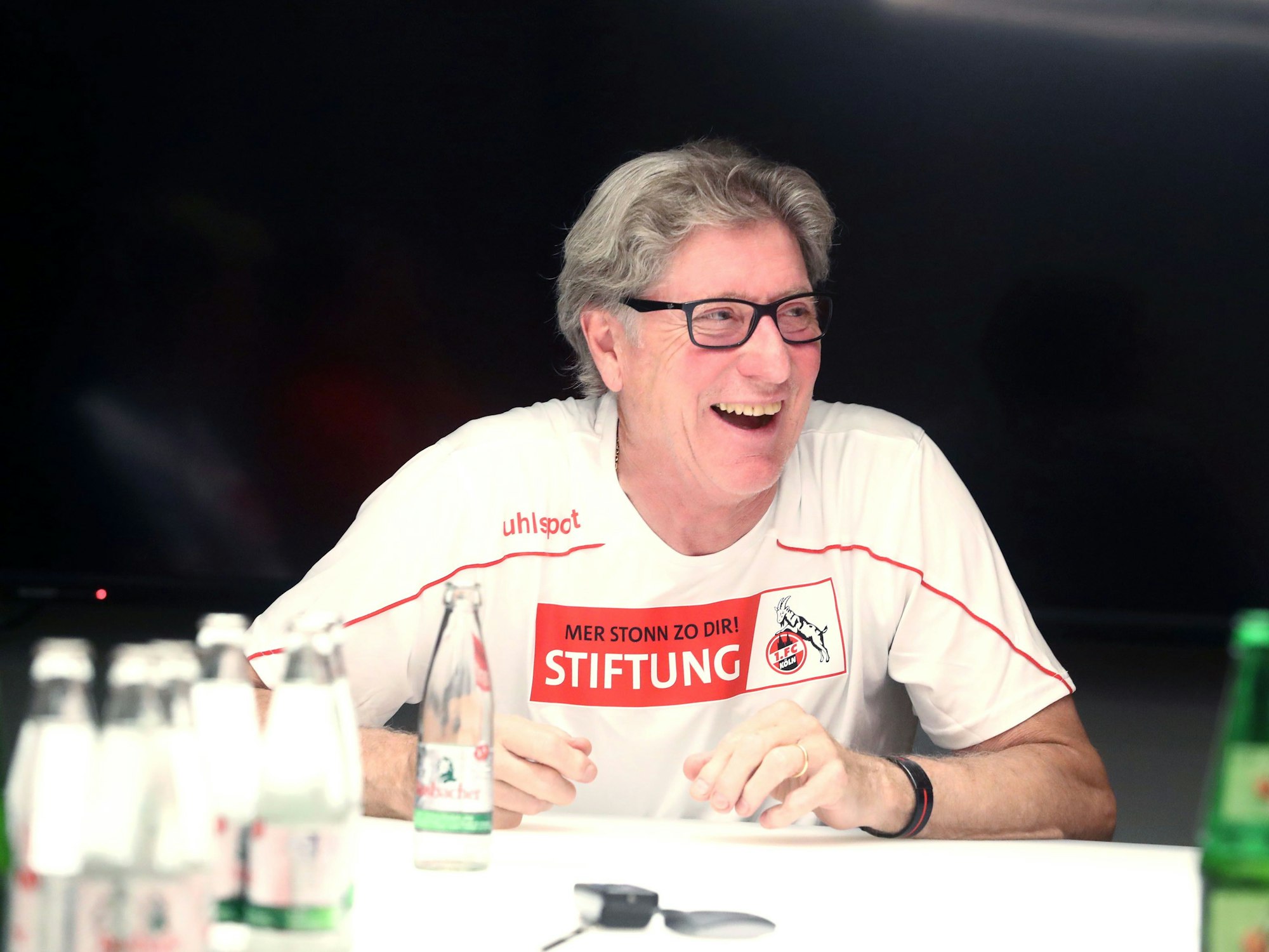 Toni Schumacher lacht, während er an einem Tisch sitzt.