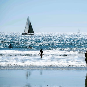 Das undatierte Symbolfoto zeigt einen Strand und das dahinterliegende Meer mit mehreren Menschen und zwei Segelbooten.