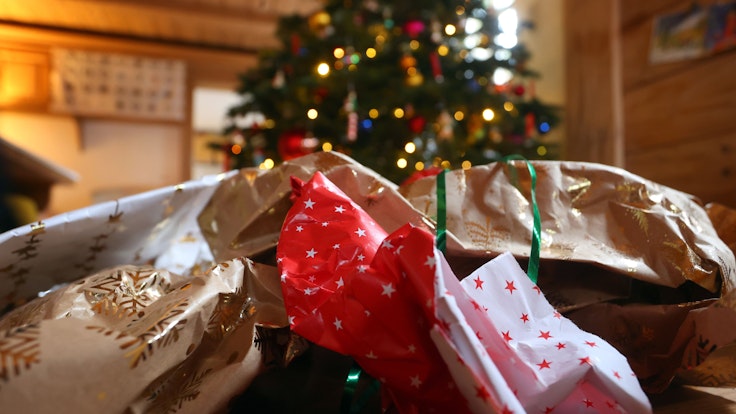 Geschenkpapier von ausgepackten Weihnachtsgeschenken liegt auf dem Boden eines Zimmers vor einem geschmücktem Weihnachtsbaum.