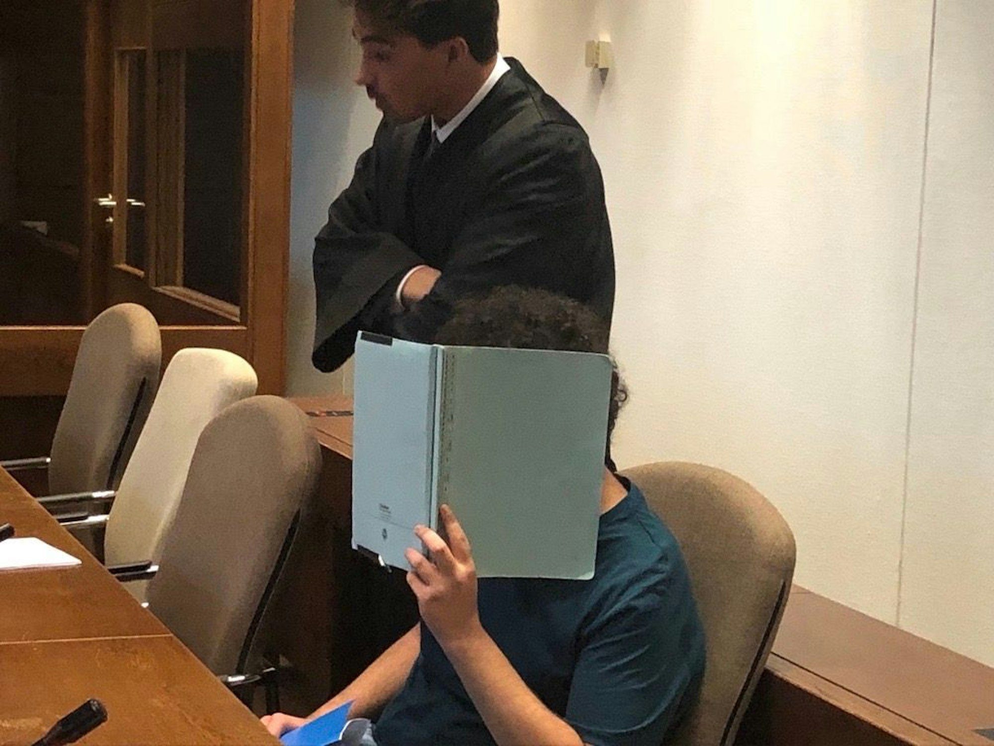 Ein Angeklagter vor Gericht hält sich einen Ordner vor das Gesicht, um auf Fotos nicht erkennbar zu sein.