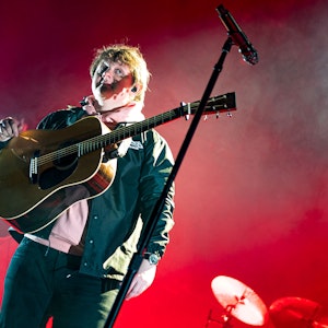 Lewis Capaldi vor einem roten Hintergrund und mit Gitarre bei einem Konzert in Köln.