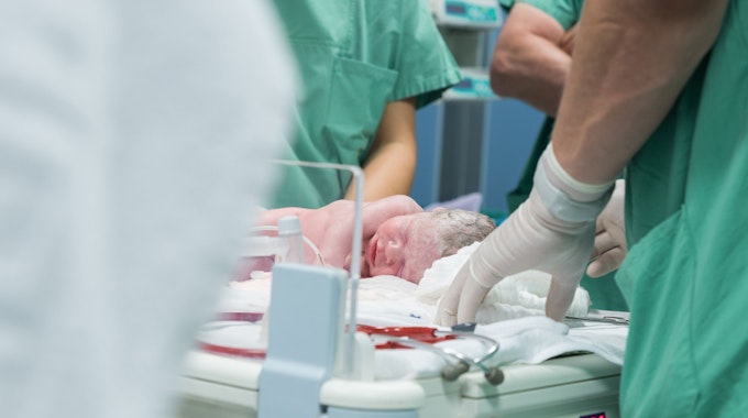 Das Symbolfoto aus dem Jahr 2016 zeigt ein Neugeborenes, das von Krankenhauspersonal in grünen Kitteln umgeben ist.