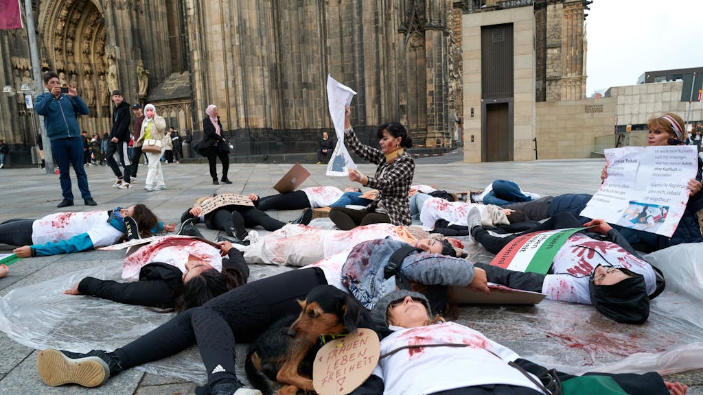 Rund 20 Personen (überwiegend Frauen) liegen in Köln auf der Domplatte – ihre T-Shirts sind mit Kunstblut beschmiert.