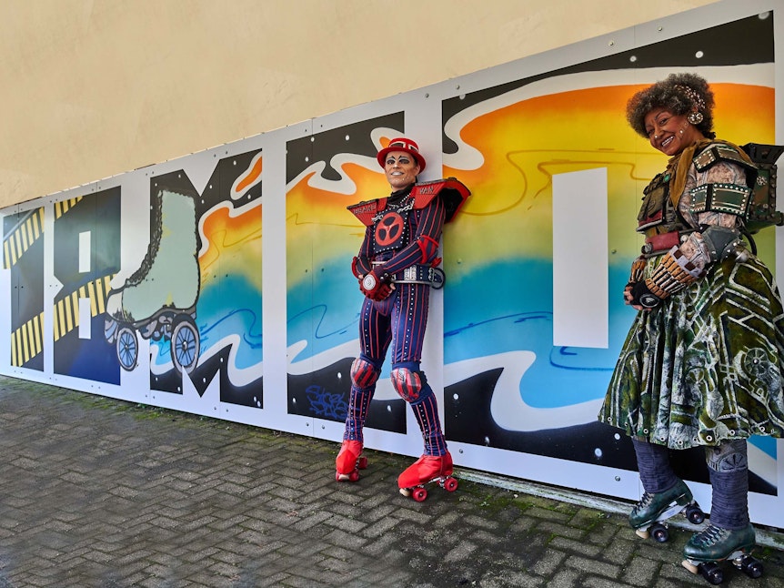 Zwei Menschen stehen vor einer Wand, auf der mit Graffiti 18 Mio. geschrieben wurde.