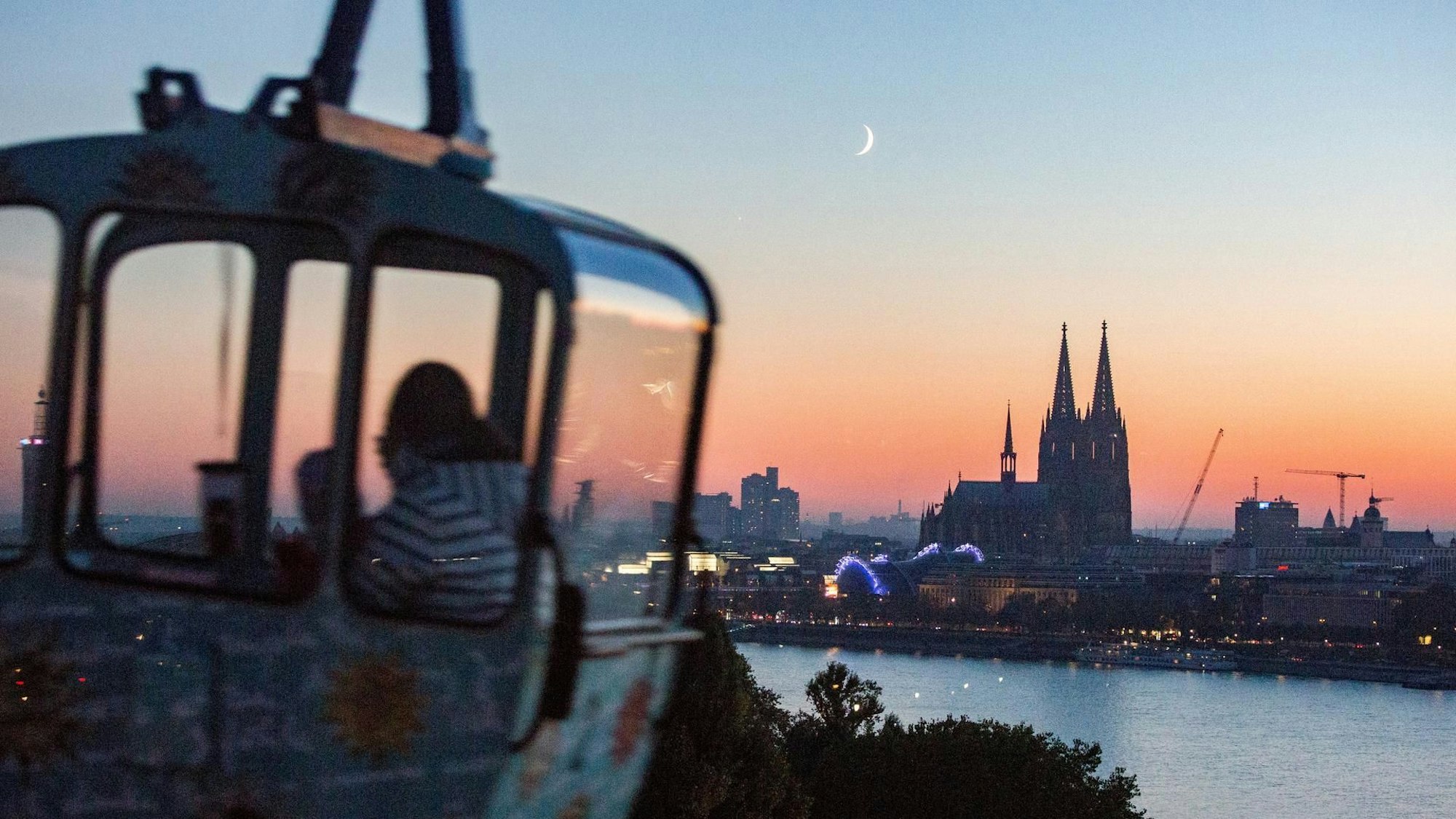 Eine Gondel der Kölner Seilbahn fährt über den Rhein mit Blick auf den Dom am Abend.