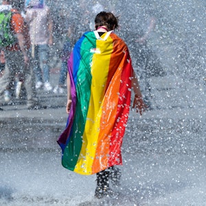 Ein Teilnehmer des Christopher Street Days (CSD) in der Innenstadt läuft bei sommerlichen Temperaturen mit einer Regenbogenfahne durch einen Springbrunnen.
