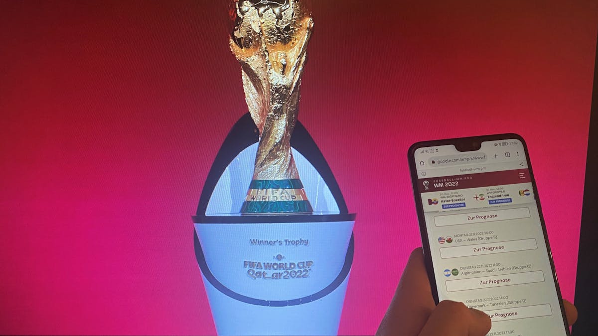 Ein Handy mit einem geöffneten Tippspiel, im Hintergrund der Pokal der Fußball-WM.