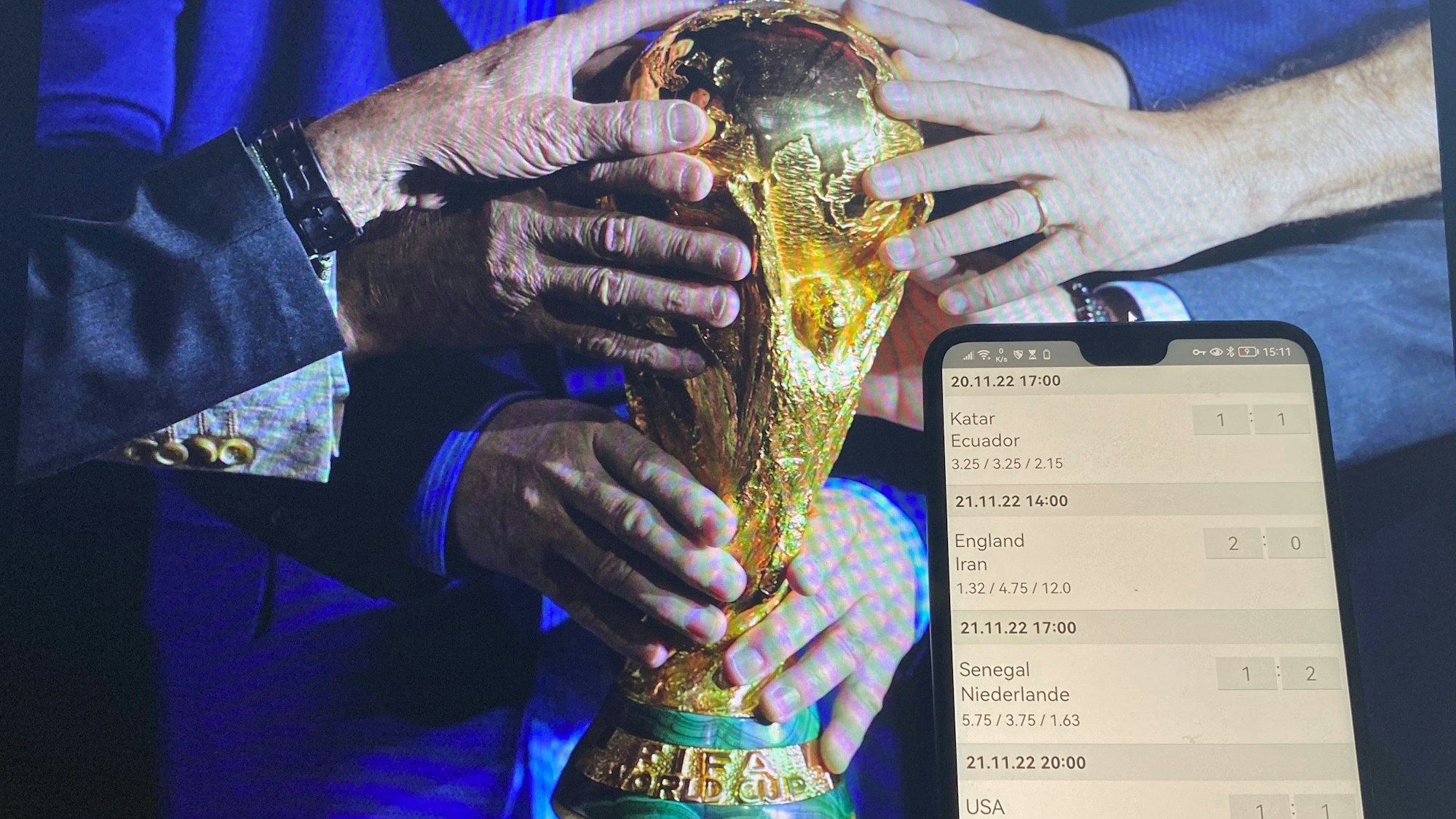 Im Hintergrund halten viele Menschen ihre Hände an den WM-Pokal, im Vordergrund ist auf einem Handy eine Tippspiel-App geöffnet