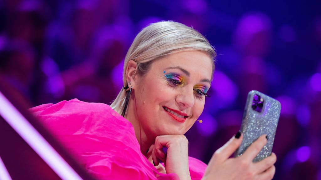 Ruth Moschner aus dem Rateteam sitzt am Ratepult im Finale der ProSieben-Show „The Masked Singer“ und blickt auf ihr Handy.
