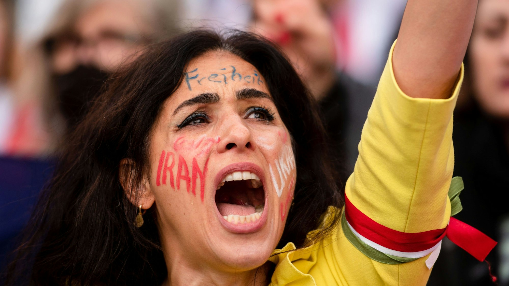 Eine Frau nahm am ·Marsch für die Freiheit·in Köln teil. Ihr Arm ist in die Luft gereckt, auf ihrer Wange steht „Iran“, auf der Stirn „Freiheit“.