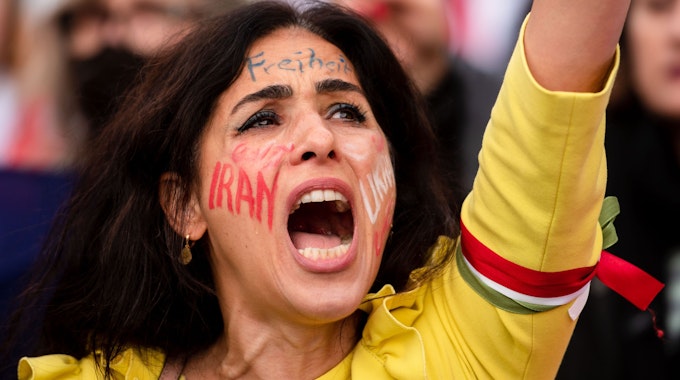 Eine Frau nahm am ·Marsch für die Freiheit·in Köln teil. Ihr Arm ist in die Luft gereckt, auf ihrer Wange steht „Iran“, auf der Stirn „Freiheit“.&nbsp;