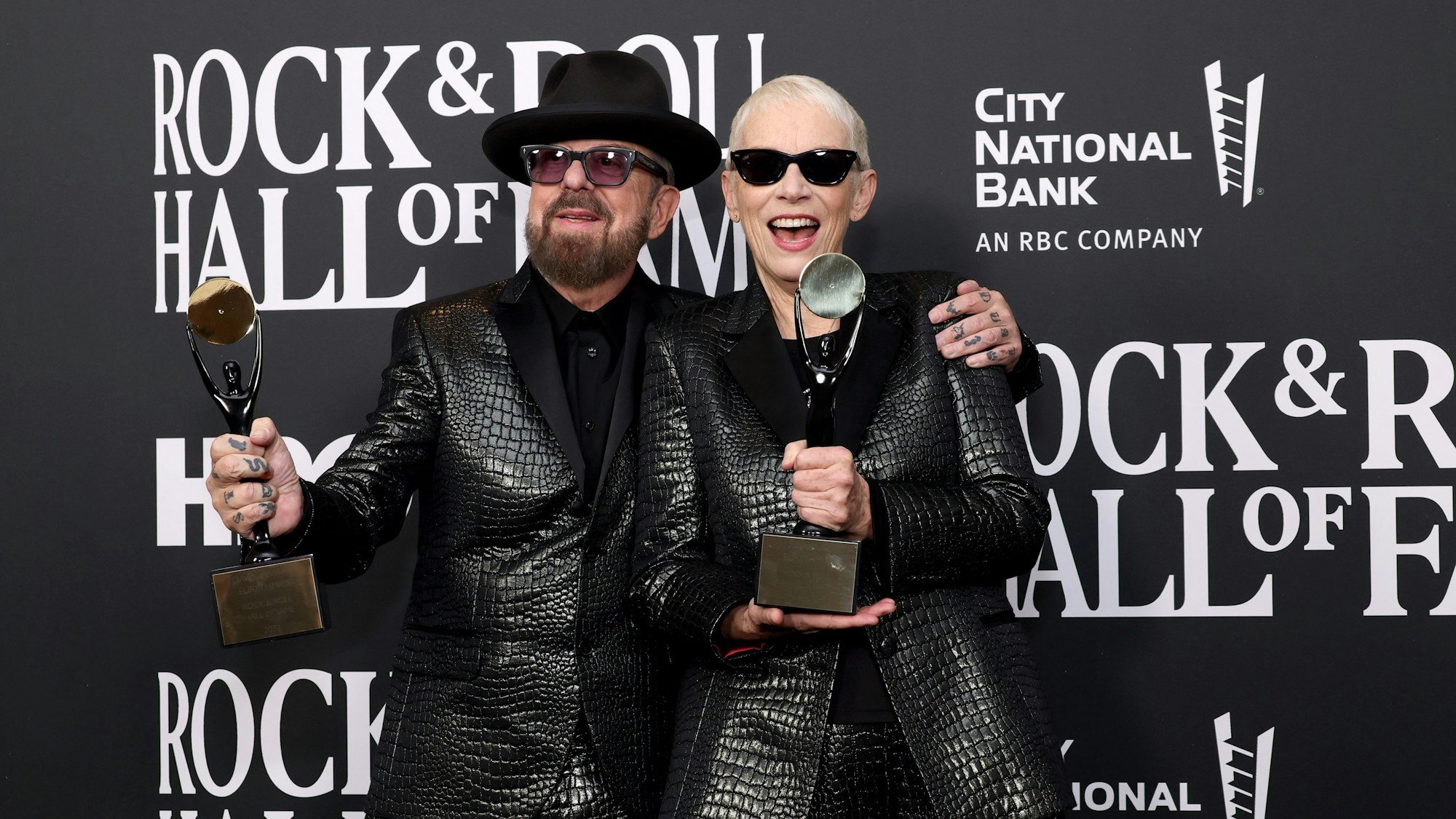 Dave Stewart und Annie Lennox bei der Pressekonferenz mit den Statuen der „Rock and Roll Hall of Fame“. Sie tragen schwarze Anzüge.
