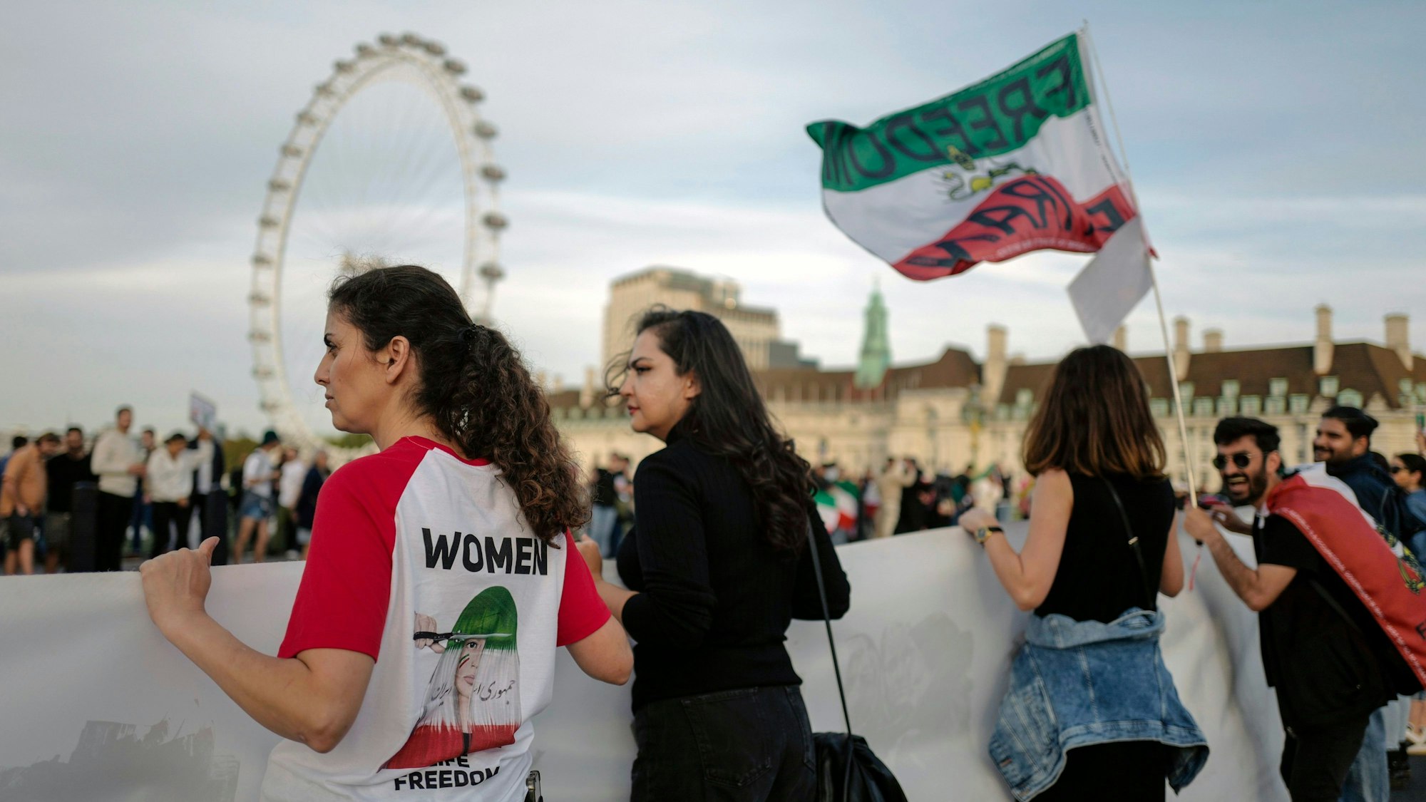 Demonstrierende halten in London Banner und Iran-Flaggen, um sich mit den Frauen im Iran zu solidarisieren. Auf dem T-Shirt einer Frau steht „Women - Freedom“. Im Hintergrund ist das London-Eye zu sehen.