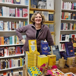 Inhaberin Ute Hentschel steht in ihrer Buchhandlung, umgeben von gut gefüllten Bücherregalen und einem Auslagetisch vor ihr.