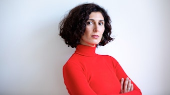 Nava Ebrahimi steht mit verschränkten Armen vor einer weißen Wand. Sie trägt einen roten Rollkragenpullover.