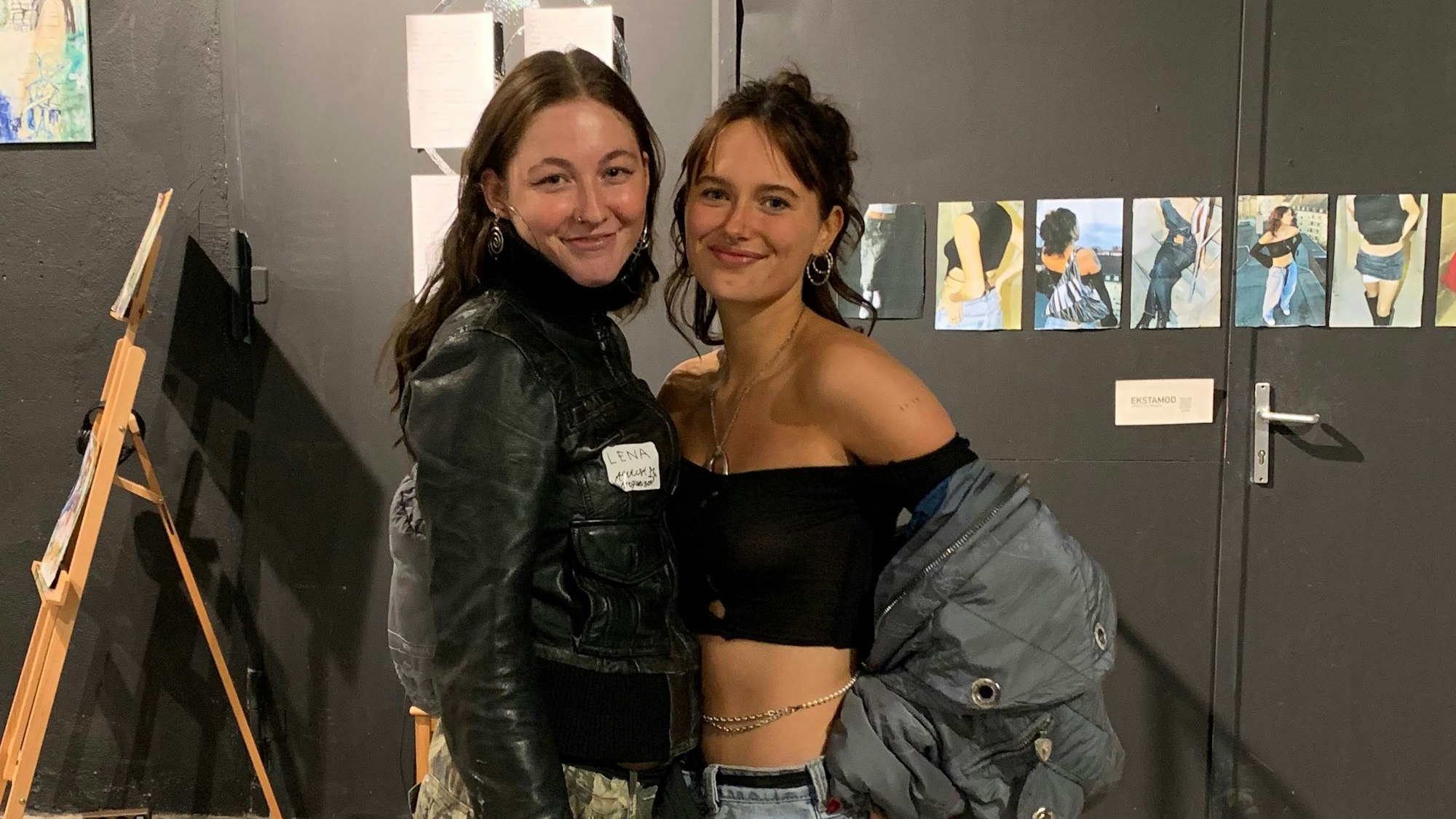 Lena Kroj und Anouk Singer stehen vor einer dunklen Wand mit kleinen Bildern aus ihrer eigenen Ausstellung.