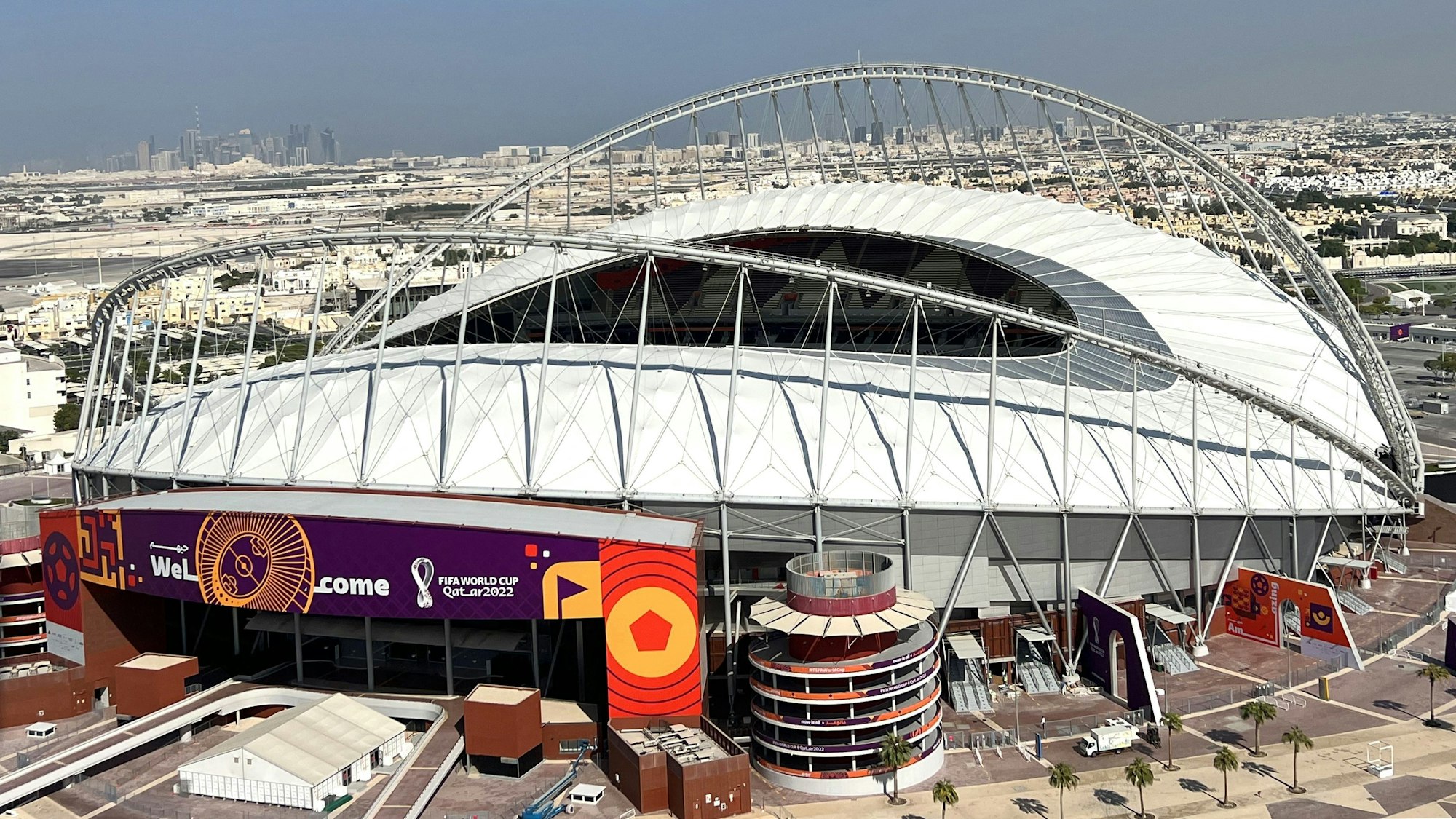 Das Khalifa Stadion in Doha ist ein ovales Gebäude mit in der Mitte offenem Dach und aufwändiger Hängedachkonstruktion an den Seiten. Im Bildhintergrund schließt sich städtische Bebauung ans Stadion an.