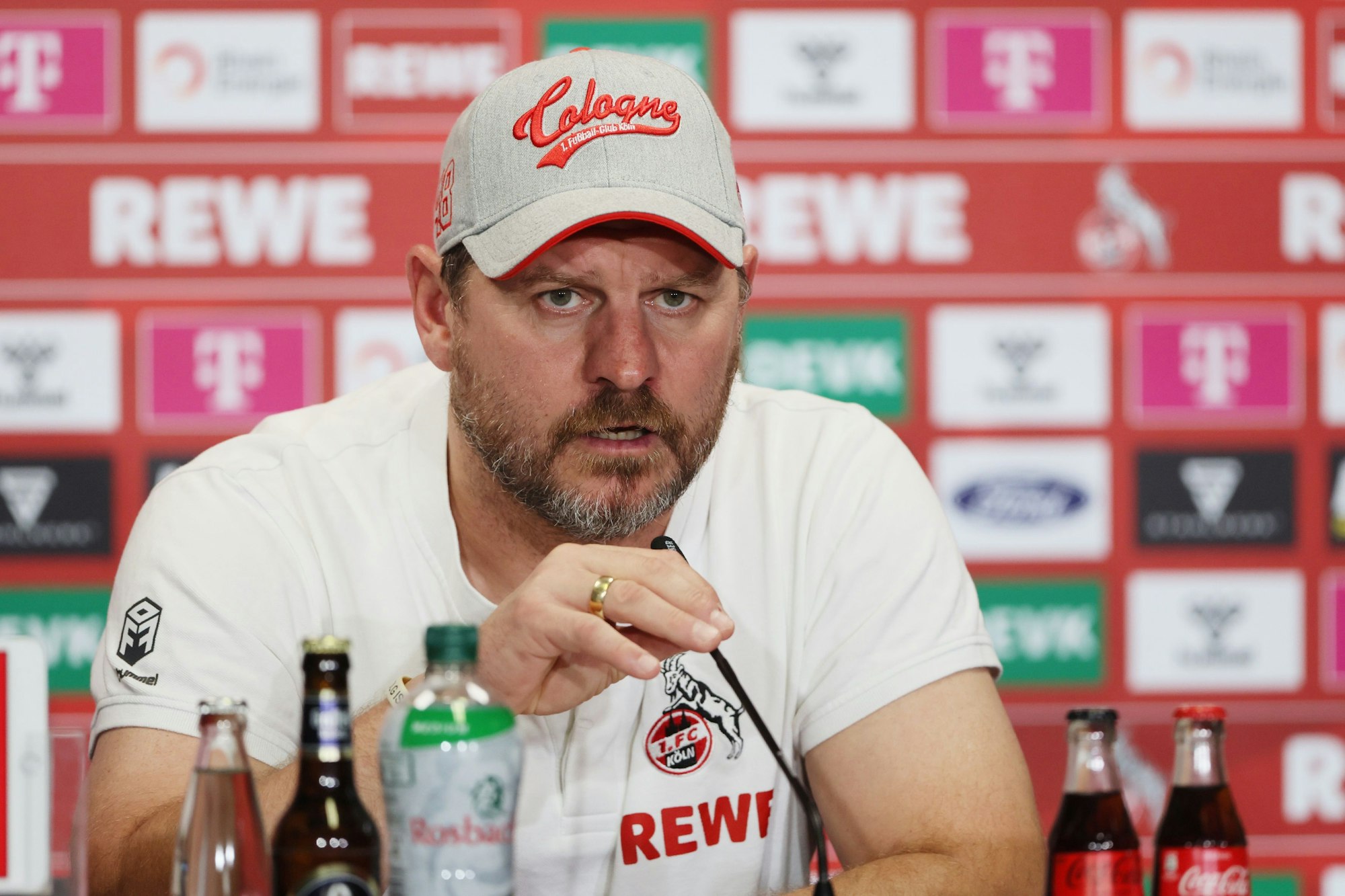 FC-Trainer Steffen Baumgart spricht auf der Pressekonferenz im Rhein-Energie-Stadion in ein Mikrofon, im Hintergrund sieht man die Sponsorenwand.