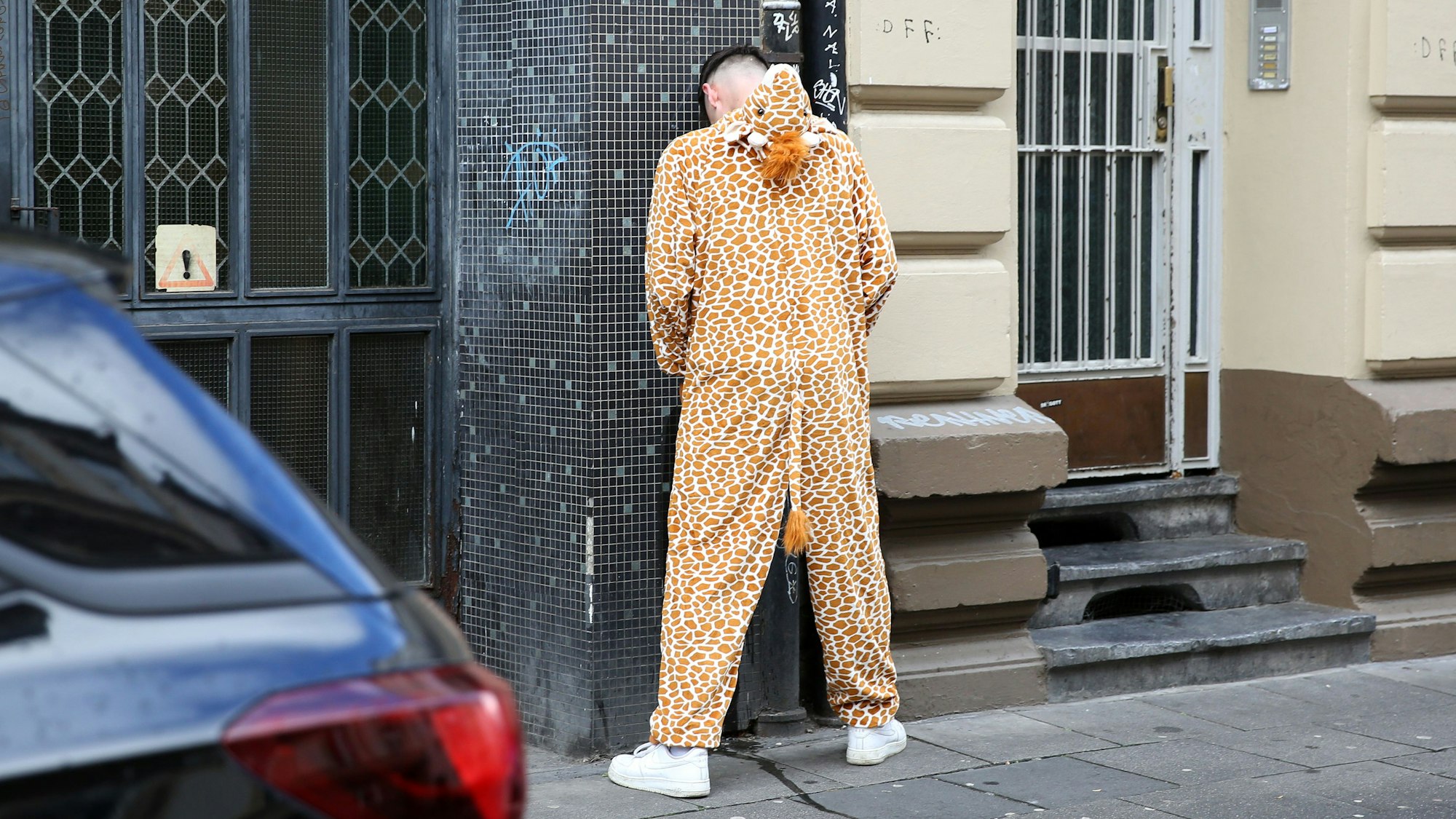 Ein Mann in einem Giraffenkostüm steht an einer Hauswand und pinkelt.