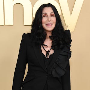 Cher lächelt für die Fotografen. Sie trägt ihre schwarzen Haare leicht gewellt und offen. Sie trägt eine schwarze Bluse und eine schwarze Kette.