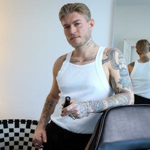 Das Bild zeigt Léon Sebastian Tiesler in einem weißen Unterhemd. Seine zahlreichen Tattoos auf dem Arm, am Brustbein, am Hals und im Gesicht sind gut zu erkennen.