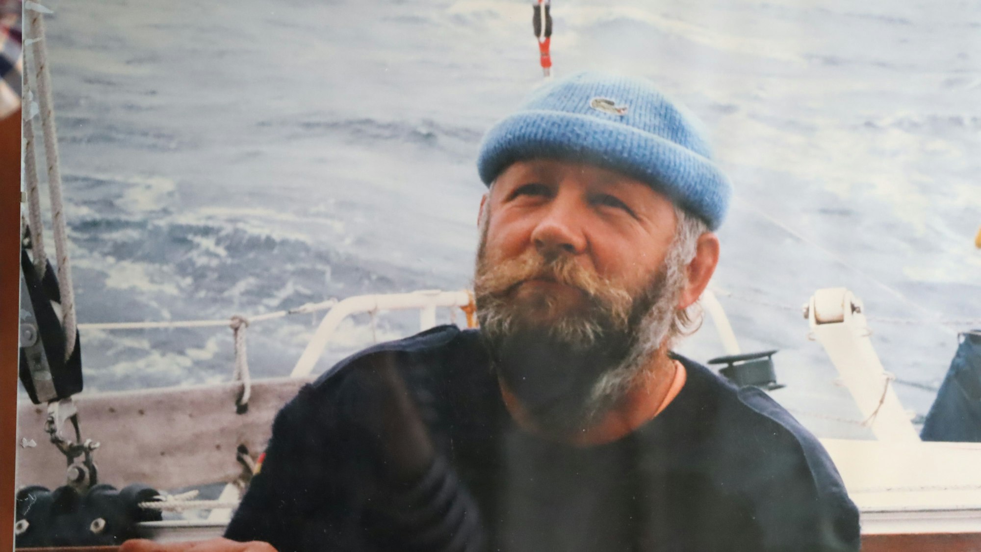 Ein Mann mit Bart, blauem Pulli und blauer Mütze sitzt auf einem Boot. Im Hintergrund das Meer.