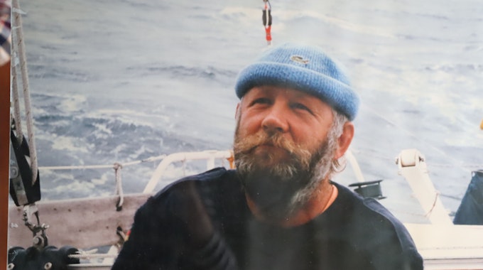 Ein Mann mit Bart, blauem Pulli und blauer Mütze sitzt auf einem Boot. Im Hintergrund das Meer.