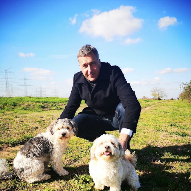 Bürgermeisterkandidat Ralph Manzke hockt mit zwei kleinen, weißen Hunden auf einer Wiese.