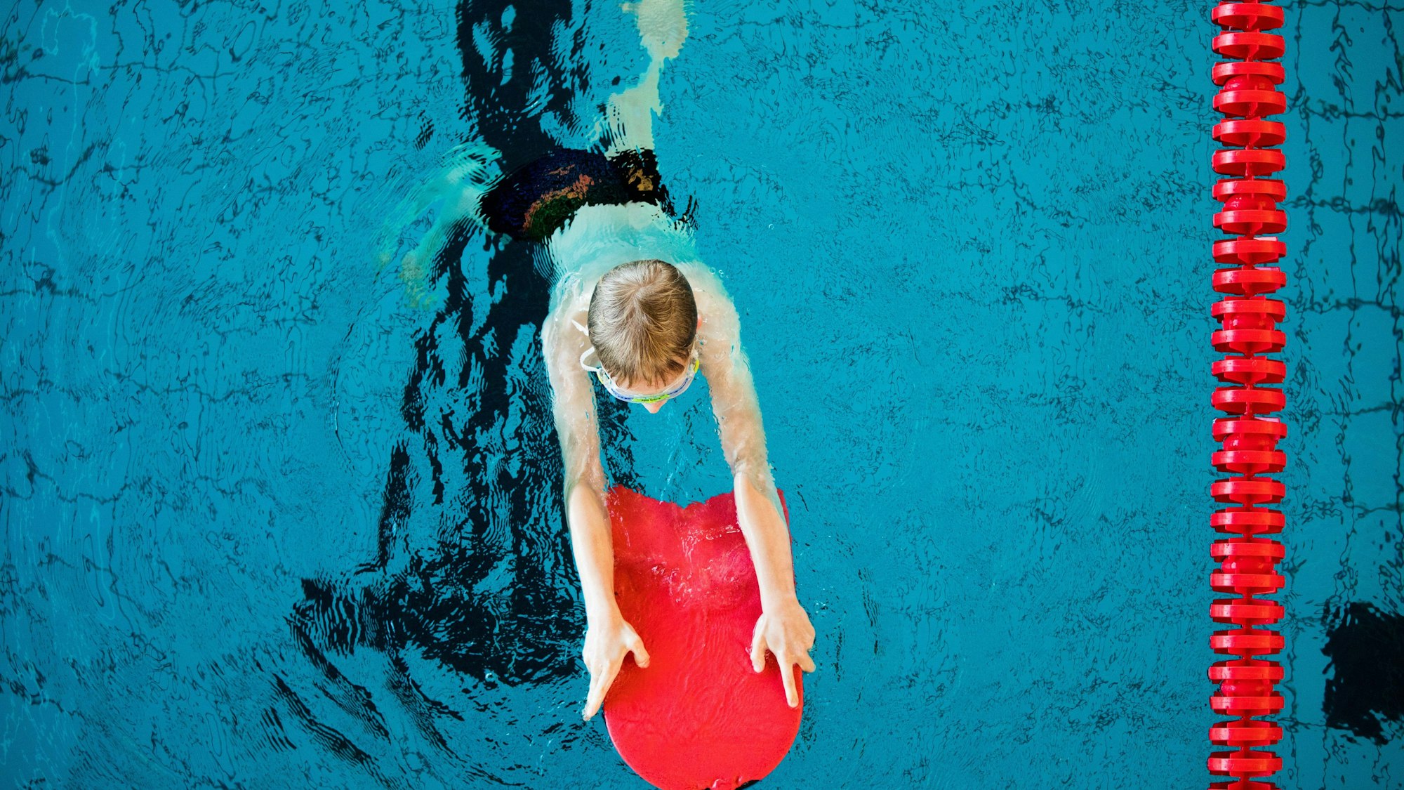 Ein Junge nimmt an einem Schwimmkurs für Kinder teil. Er hält sich mit einem roten Schwimmbrett über Wasser.